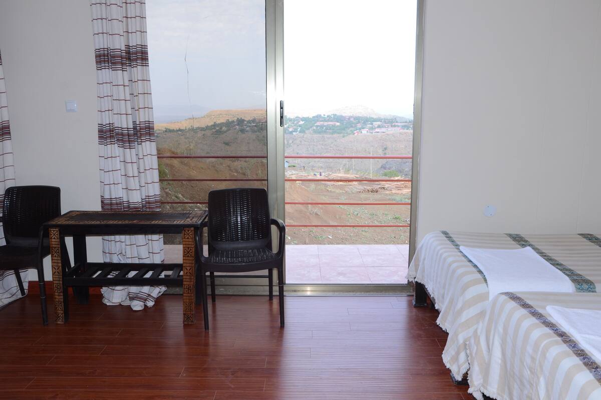 A room with a bed, a table, and a chair. There is a view of a landscape through a large window.