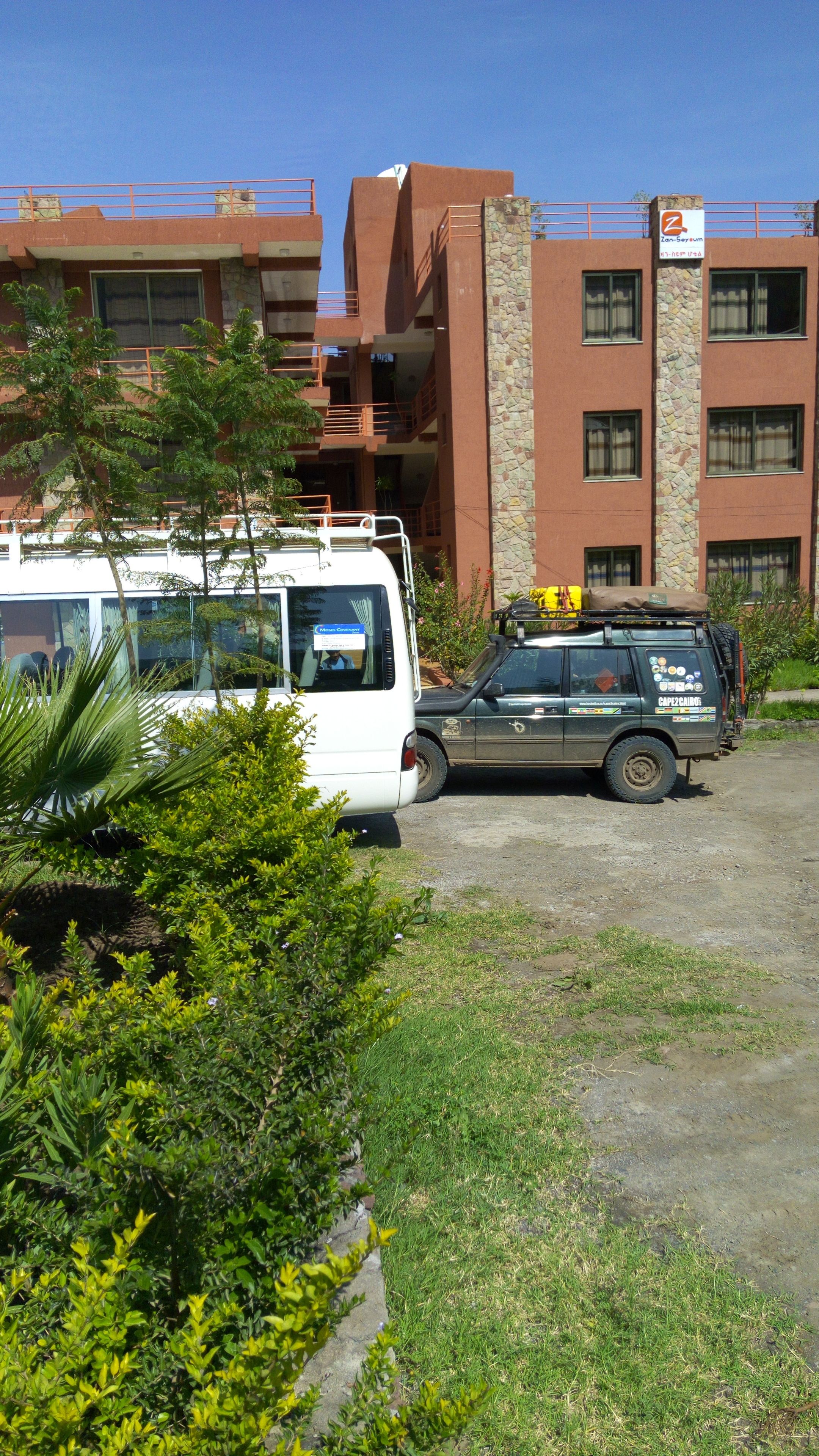 Un espace de stationnement où se trouvent un fourgon blanc et un SUV vert. On aperçoit des immeubles avec des balcons et, au fond, un mur de pierre.