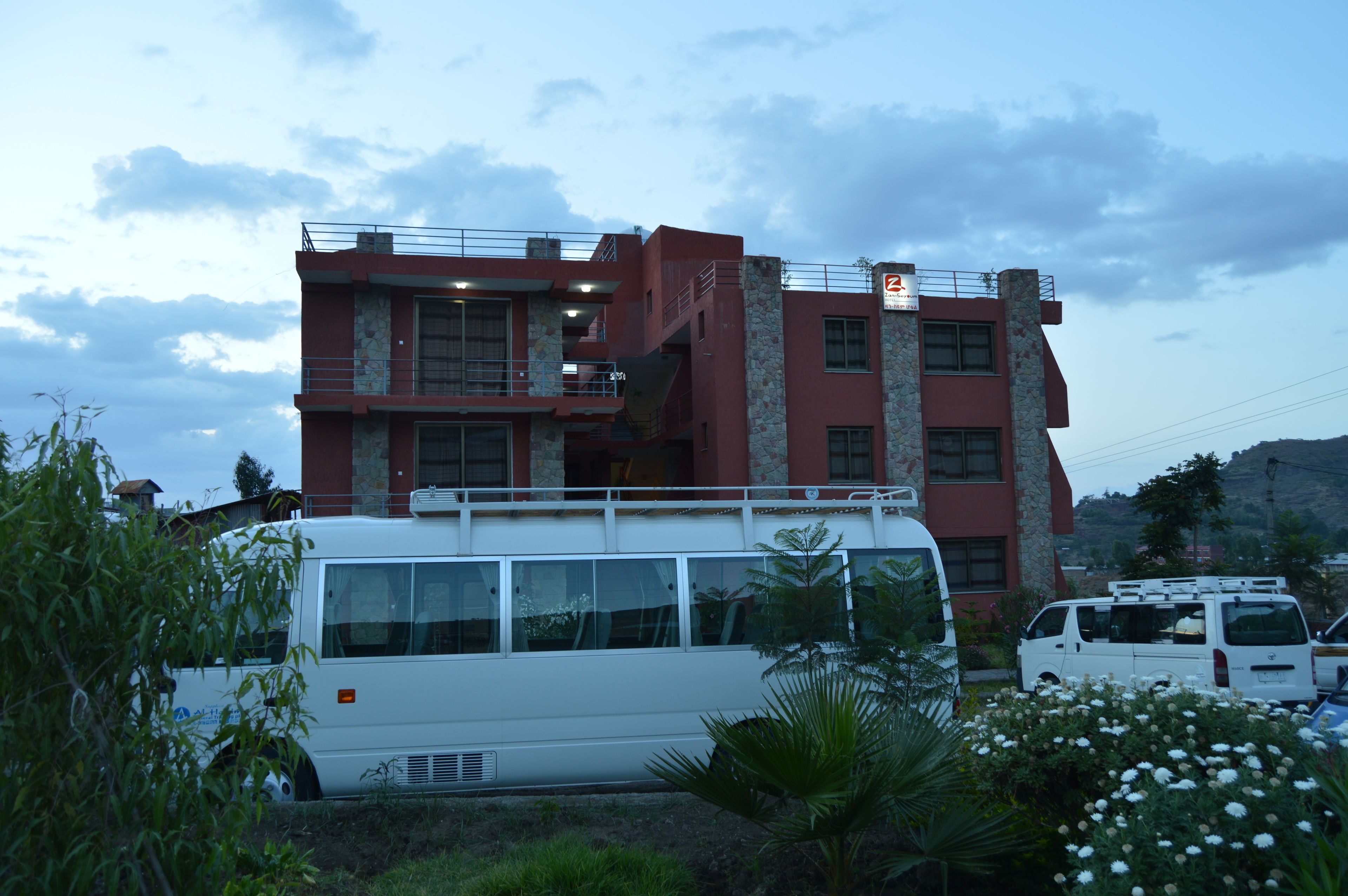 A multi-story building with a balcony on the second floor, a white van parked in front, and a mountainous backdrop.