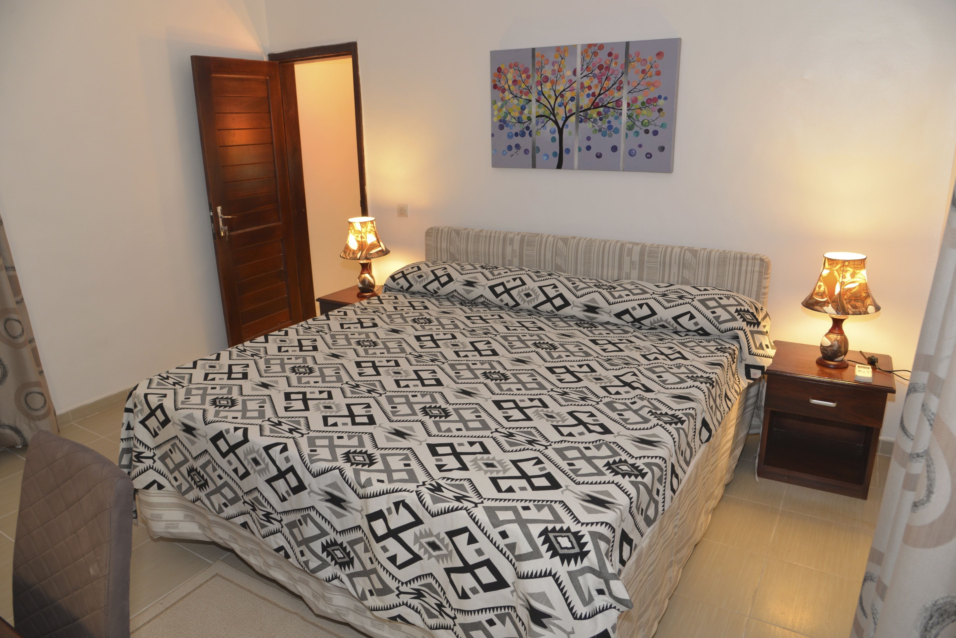 A bedroom with a patterned bedspread, two bedside tables with lamps, a door, and a wall painting.