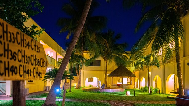 A courtyard with palm trees, a thatched-roof structure, and a building with illuminated windows. - Hotel La Coccinelle (Bamako)