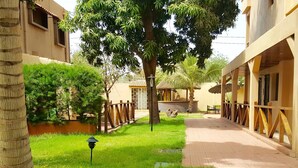 A courtyard with a pathway, palm trees, and a building with a tiled roof. - Hotel La Coccinelle (Bamako)
