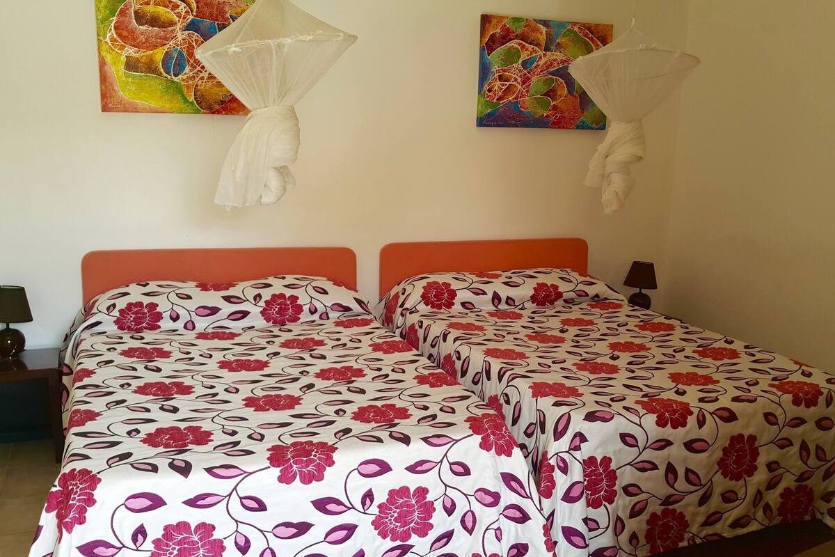 Una cama doble con ropa de cama floral, una mesita de noche con lámpara y dos mosquiteras colgadas en la pared.