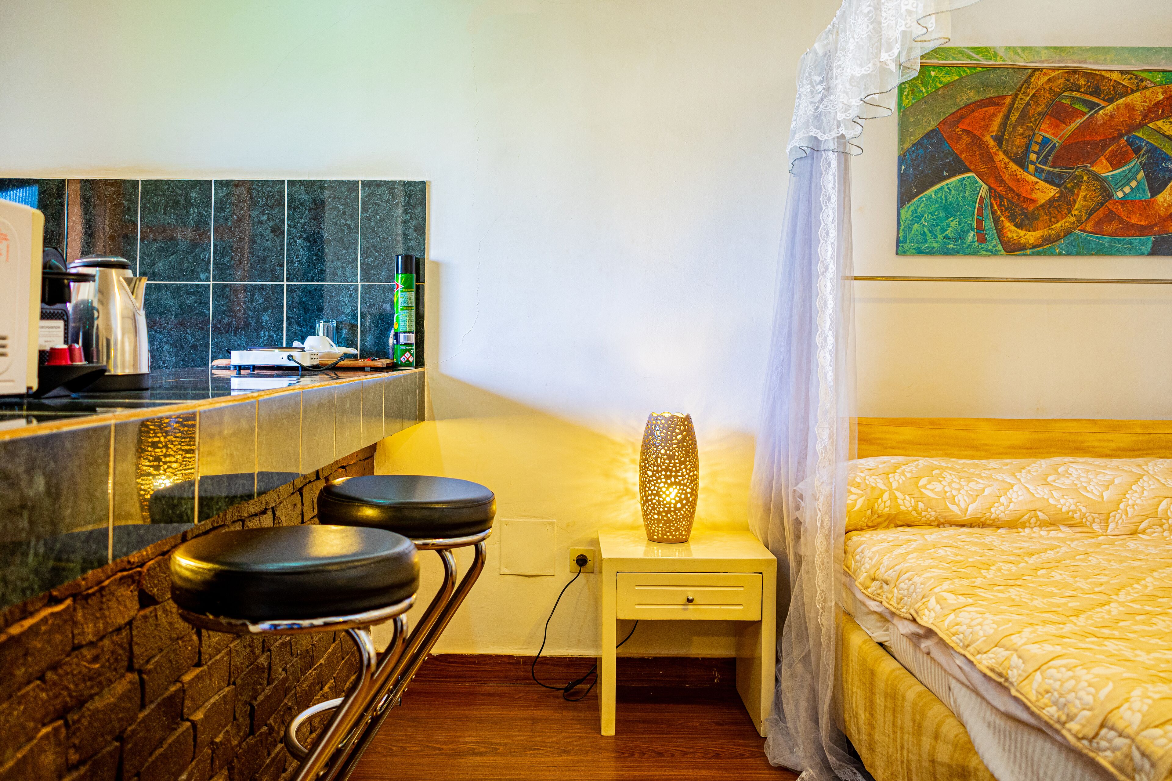 A hotel room with a bed, a nightstand with a lamp, a bar area with stools, and a tiled backsplash.