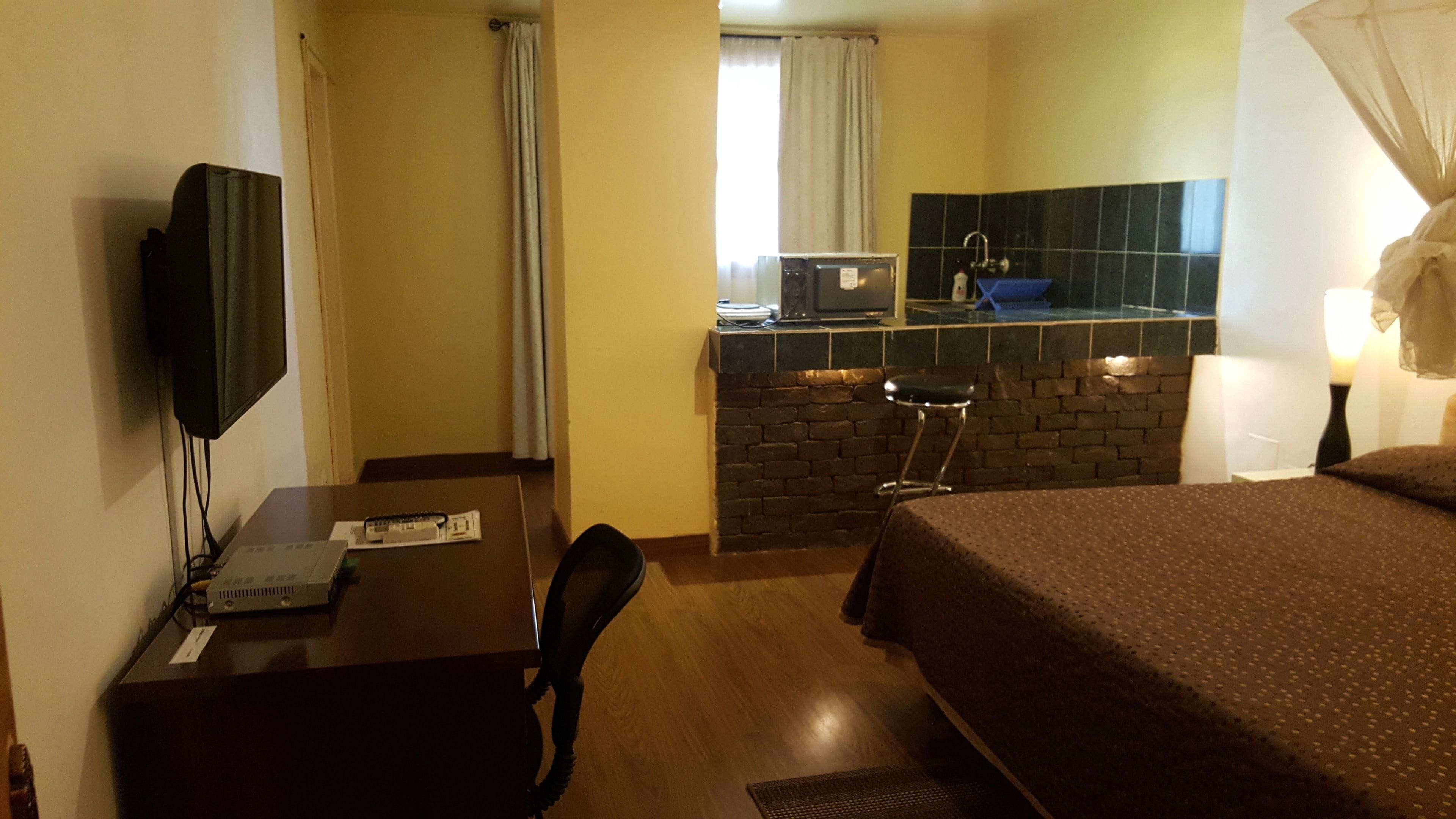 A hotel room with a bed, desk, chair, television, and a kitchenette area with a sink and stove.