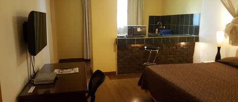 A hotel room with a bed, desk, chair, television, and a kitchenette area with a sink and stove.
