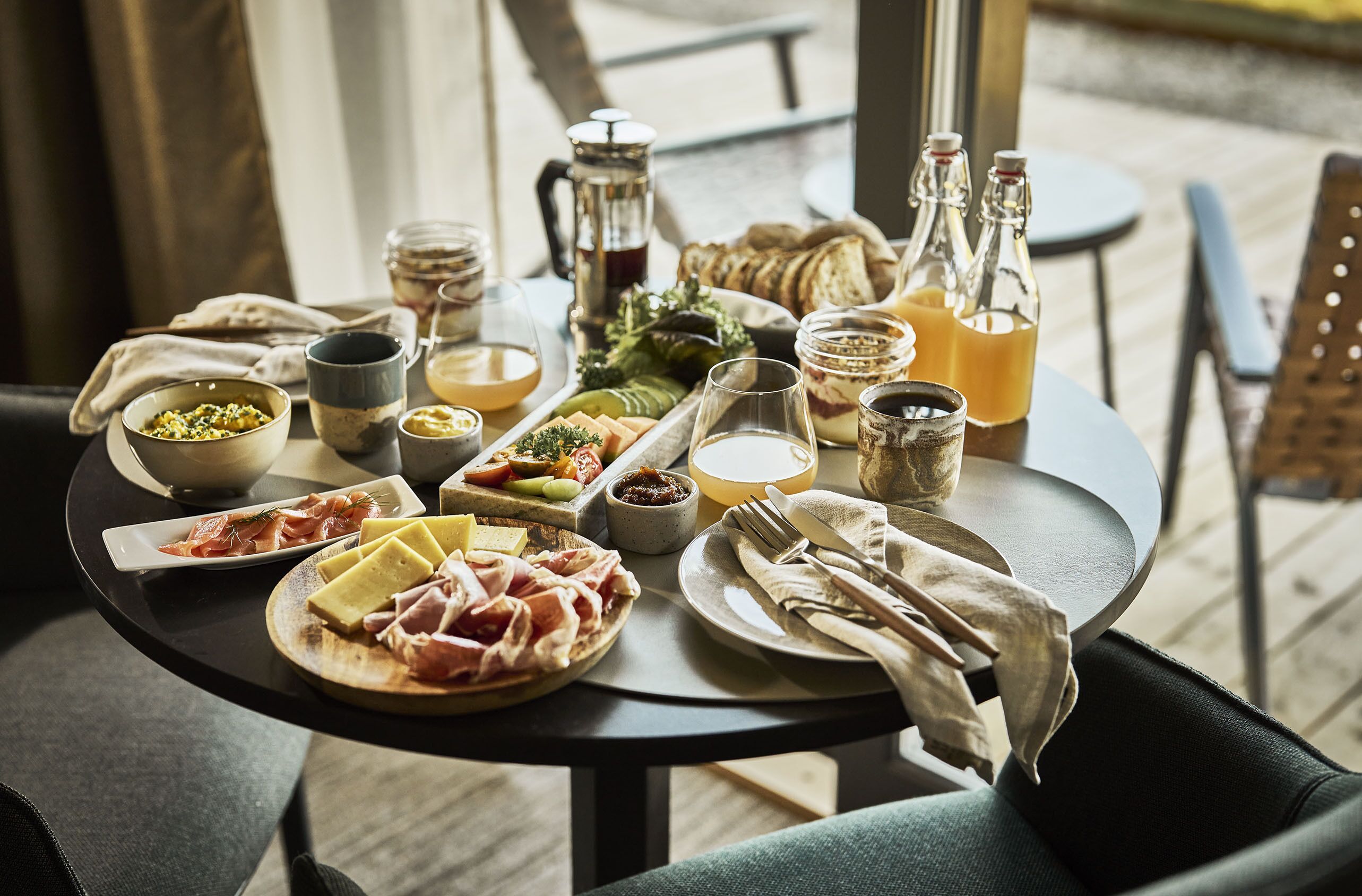 Una mesa dispuesta con diversos alimentos y bebidas, incluyendo queso, carnes y salsas, sobre una mesa de madera en una habitación bien iluminada.