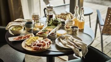 Una mesa dispuesta con diversos alimentos y bebidas, incluyendo queso, carnes y salsas, sobre una mesa de madera en una habitación bien iluminada.