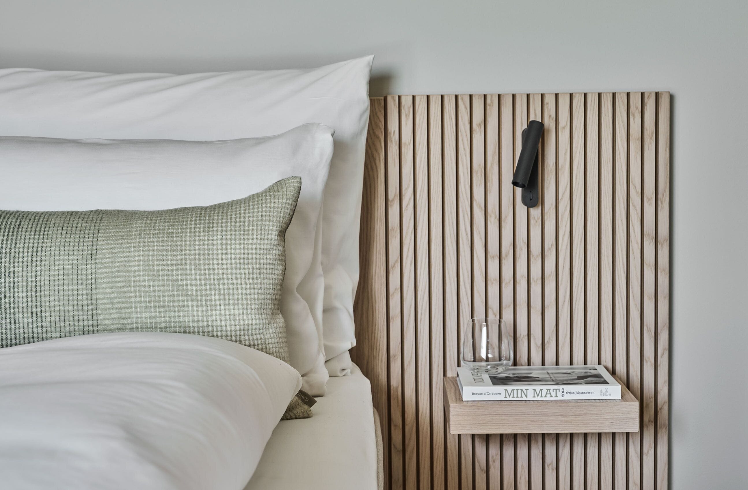 Een net opgemaakt bed met witte en geruitte kussens, een nachtkastje met een boek en een glas, en een houten wandpaneel met latten.