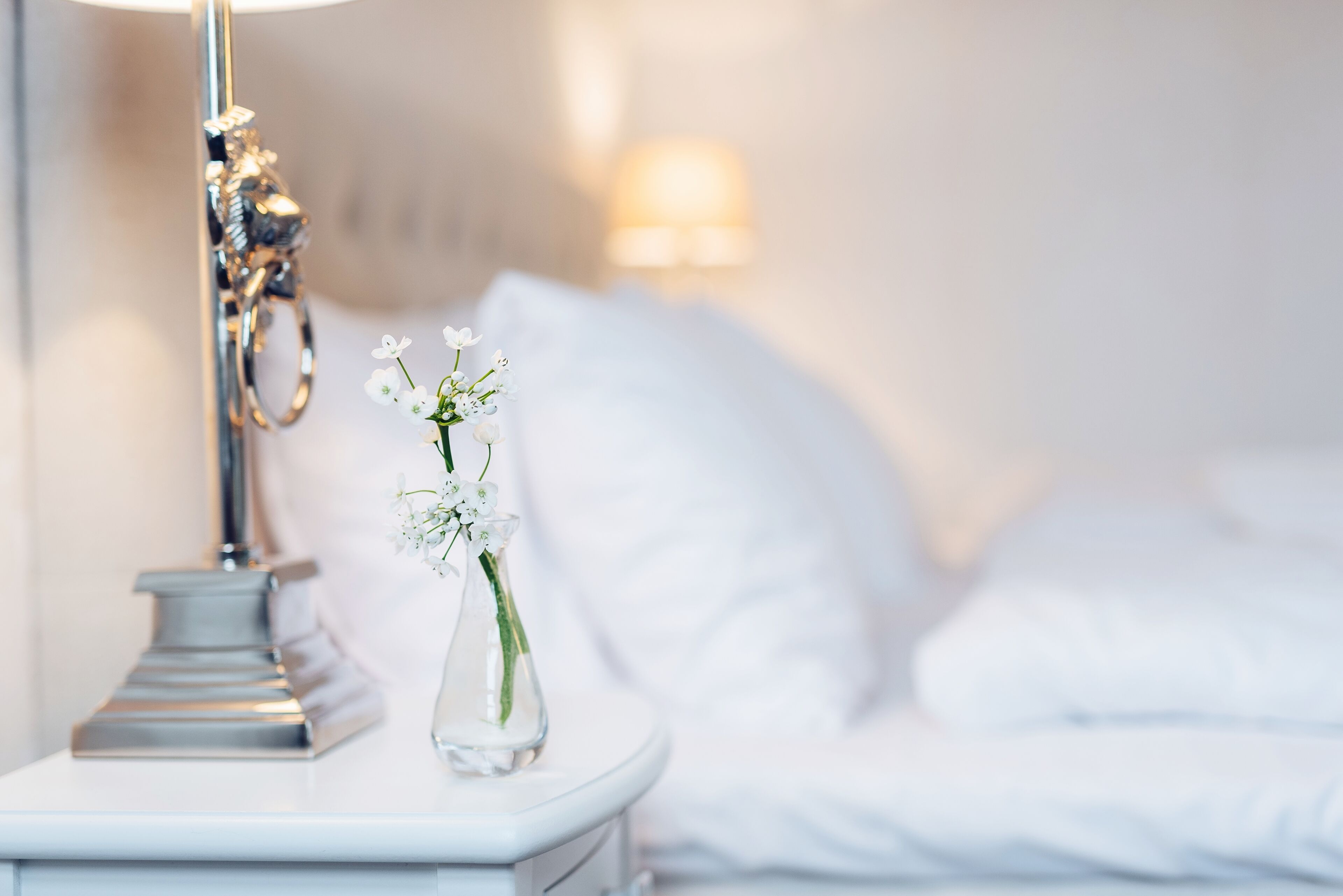 Een keurig opgemaakt bed met witte lakens, een nachtkastje met een lamp en een vaas met bloemen.