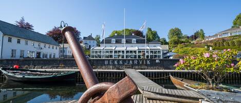 Un muelle de madera con un ancla oxidada, un barco blanco llamado 'BEKKJARVIK' amarrado al muelle, y un pequeño bote en el fondo.