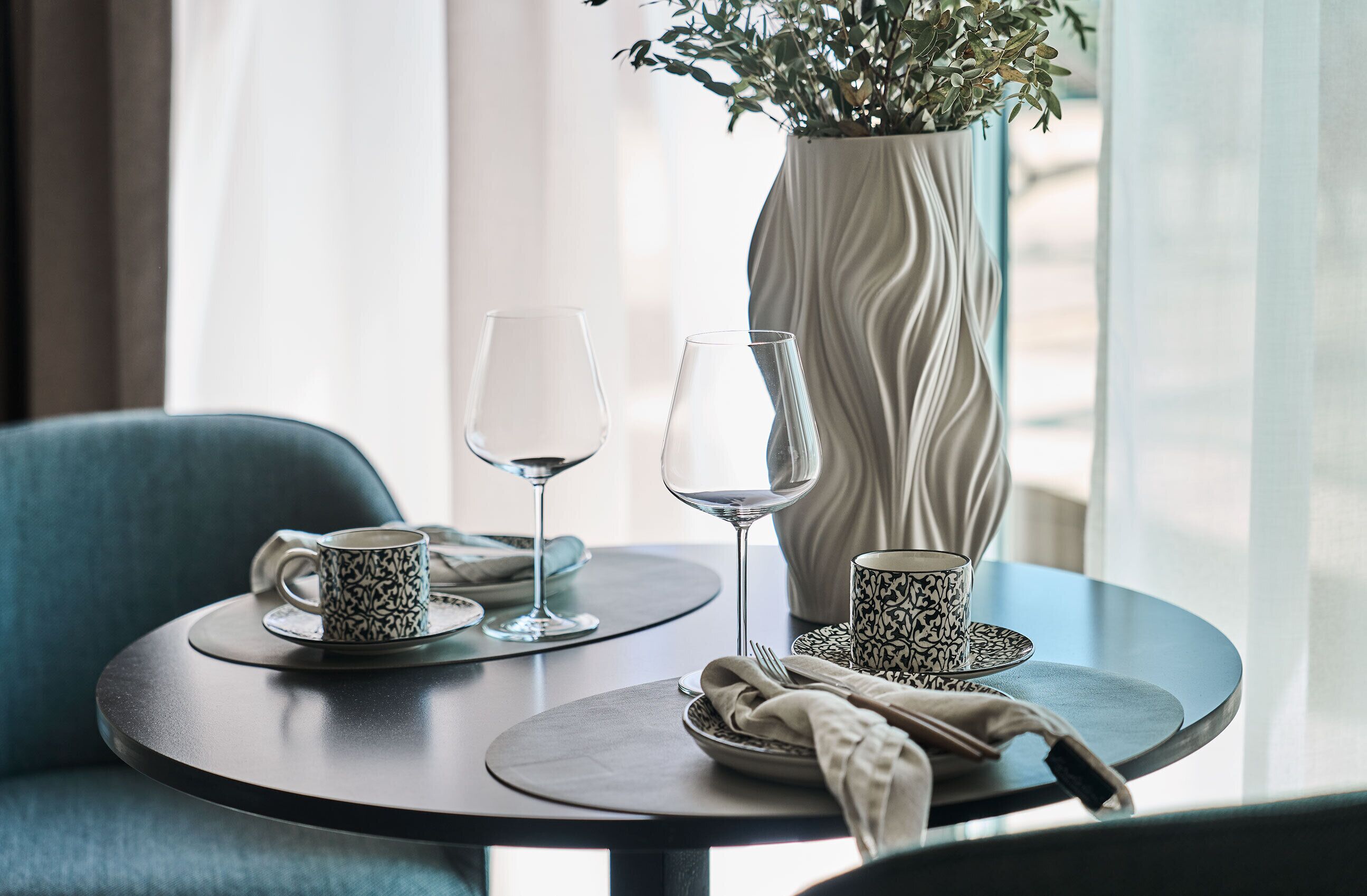 Una mesa de comedor con dos copas de vino, un florero con follaje y una taza estampada.