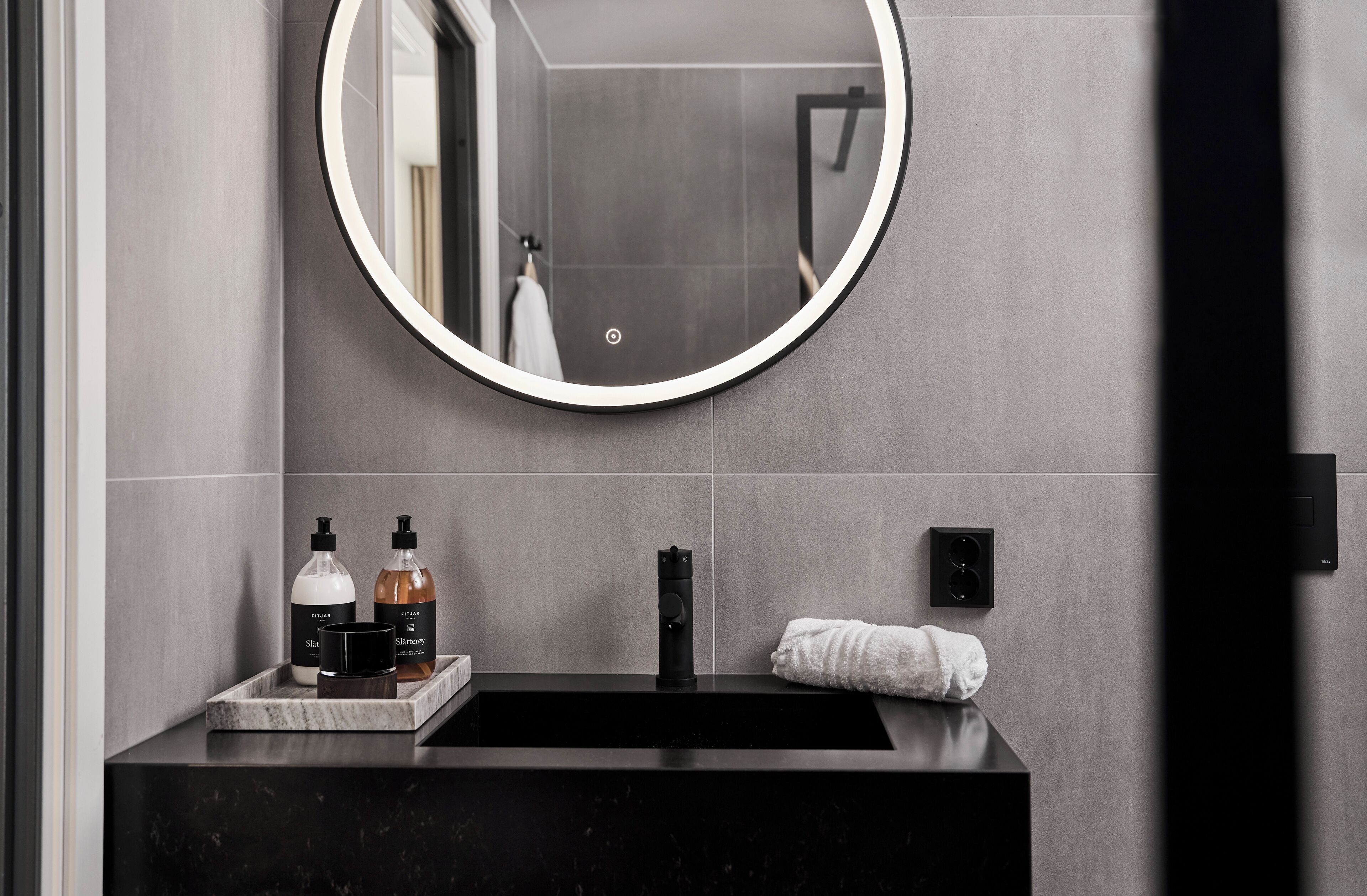 A modern bathroom with a round mirror, a black sink, and toiletries on a marble tray.