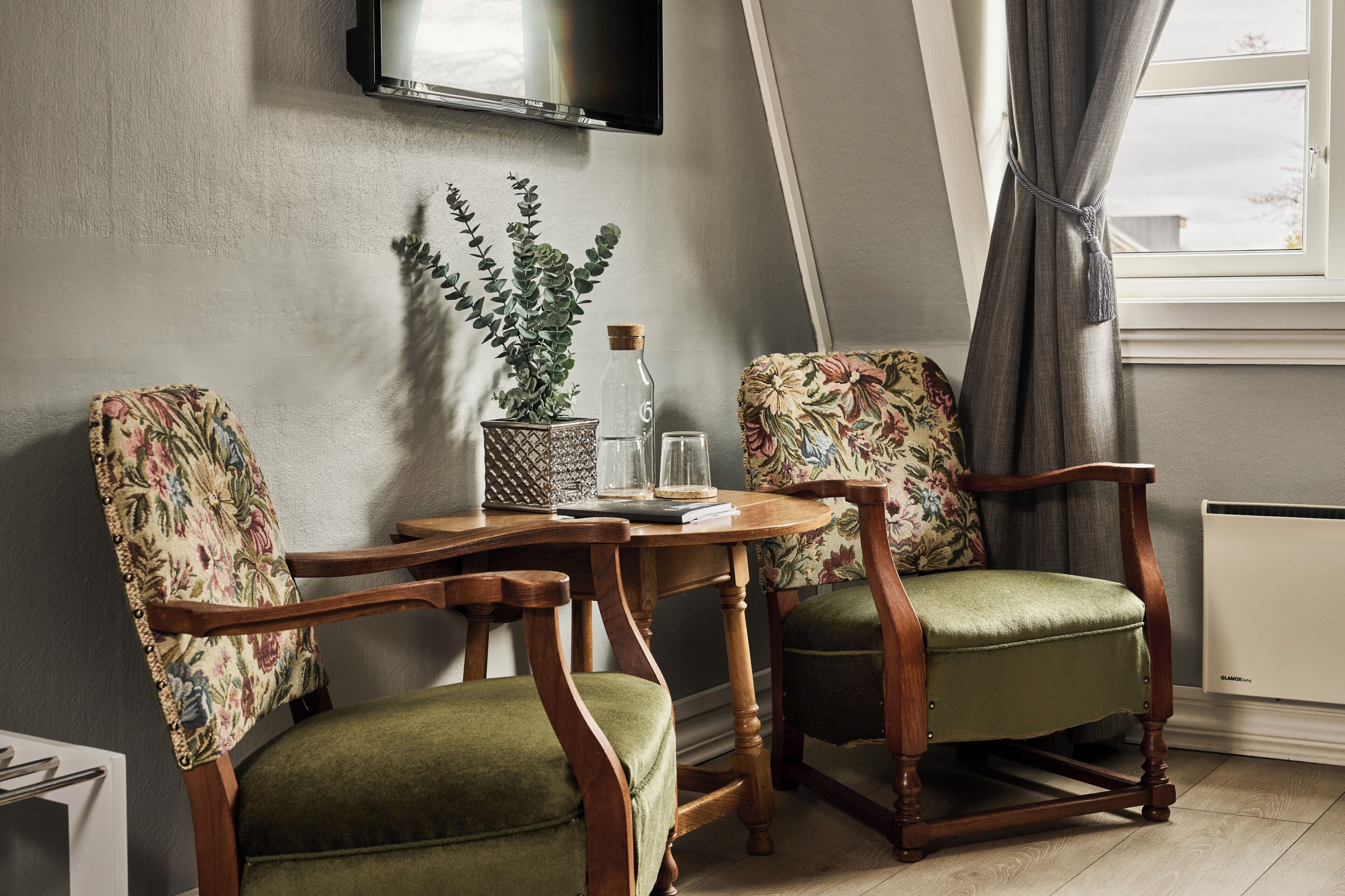 Una habitación con dos sillas de madera con tapicería floral, una mesita con una botella de vidrio y un libro, y un televisor montado en la pared.