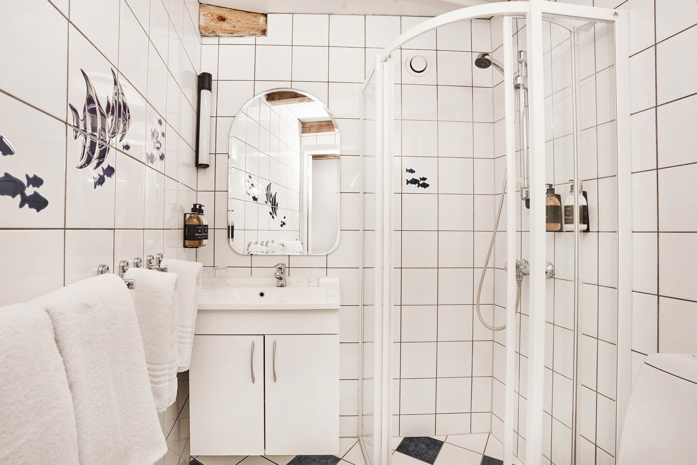 Un baño con azulejos blancos, un lavamanos blanco, un espejo y una zona de ducha.