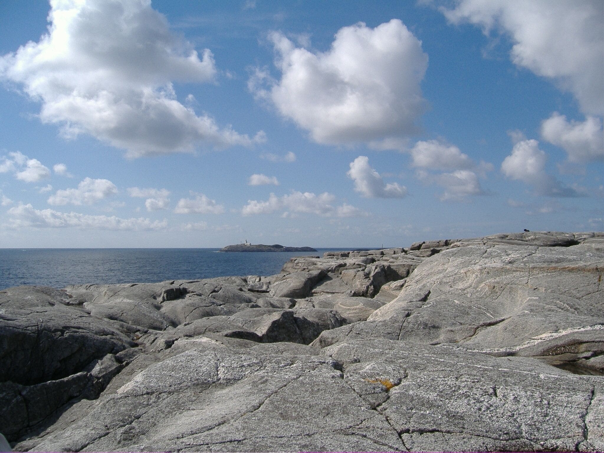 A rocky shoreline with a lighthouse in the distance under a partly cloudy sky.