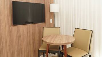 A hotel room with a flat-screen TV mounted on a wooden wall, a round table with two chairs, and a floor lamp.