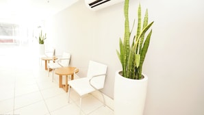 A bright, modern waiting area with white furniture, a large potted plant, and an air conditioning unit on the wall. - Promenade Fusion Itaguaí (Itaguai)