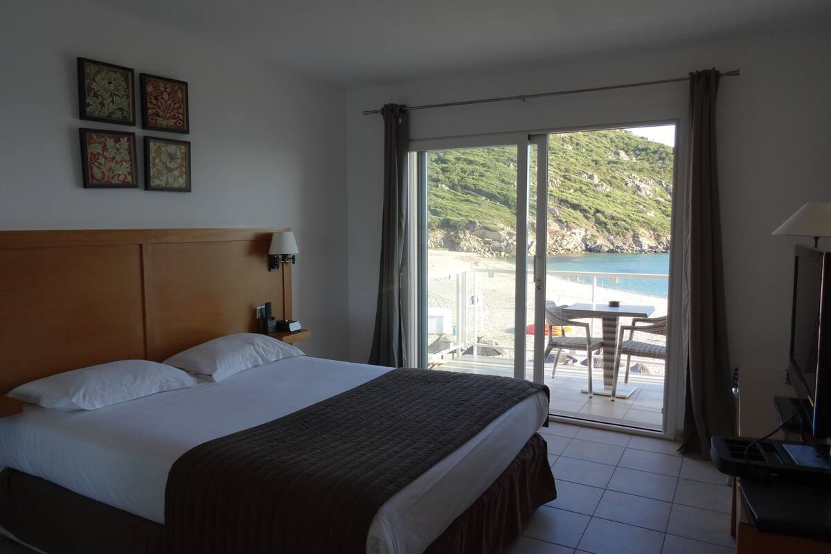 A hotel room with a bed, a desk with a computer, a chair, and a view of the beach and sea through large windows.