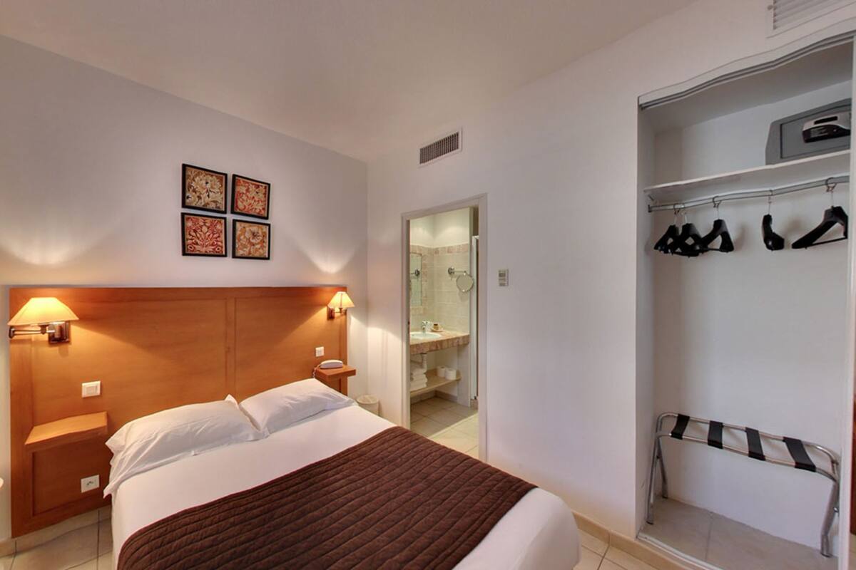 A hotel room with a bed, bedside lamps, a wooden headboard, a brown throw blanket, a closet with hanging clothes, and a bathroom with a shower and toilet.