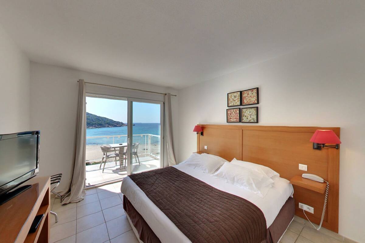 A hotel room with a bed, a TV, a phone, and a balcony with a view of the sea.
