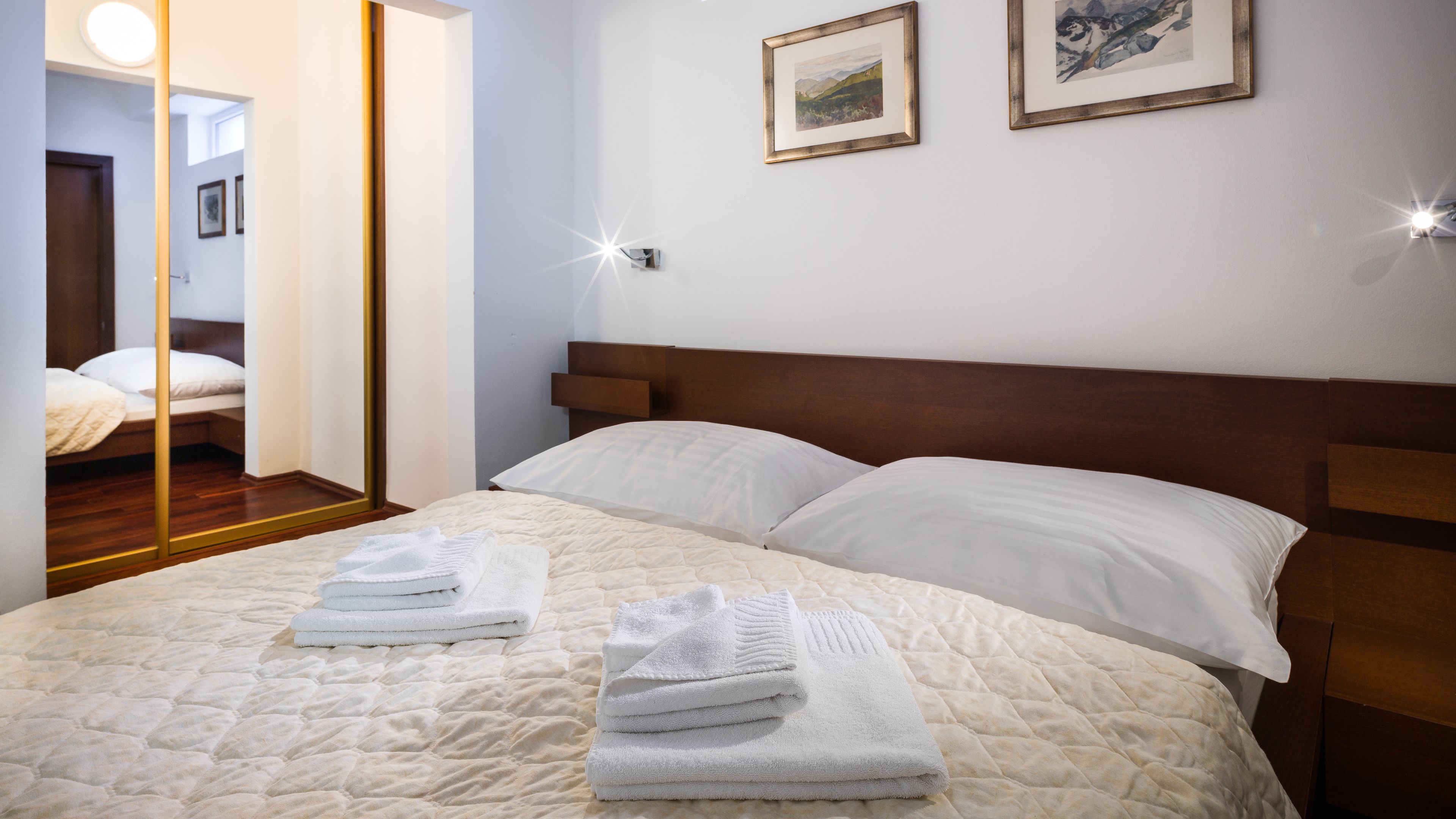 Cama bem arrumada com lençóis brancos e edredom acolchoado, cabeceira de madeira e duas fotografias emolduradas na parede.