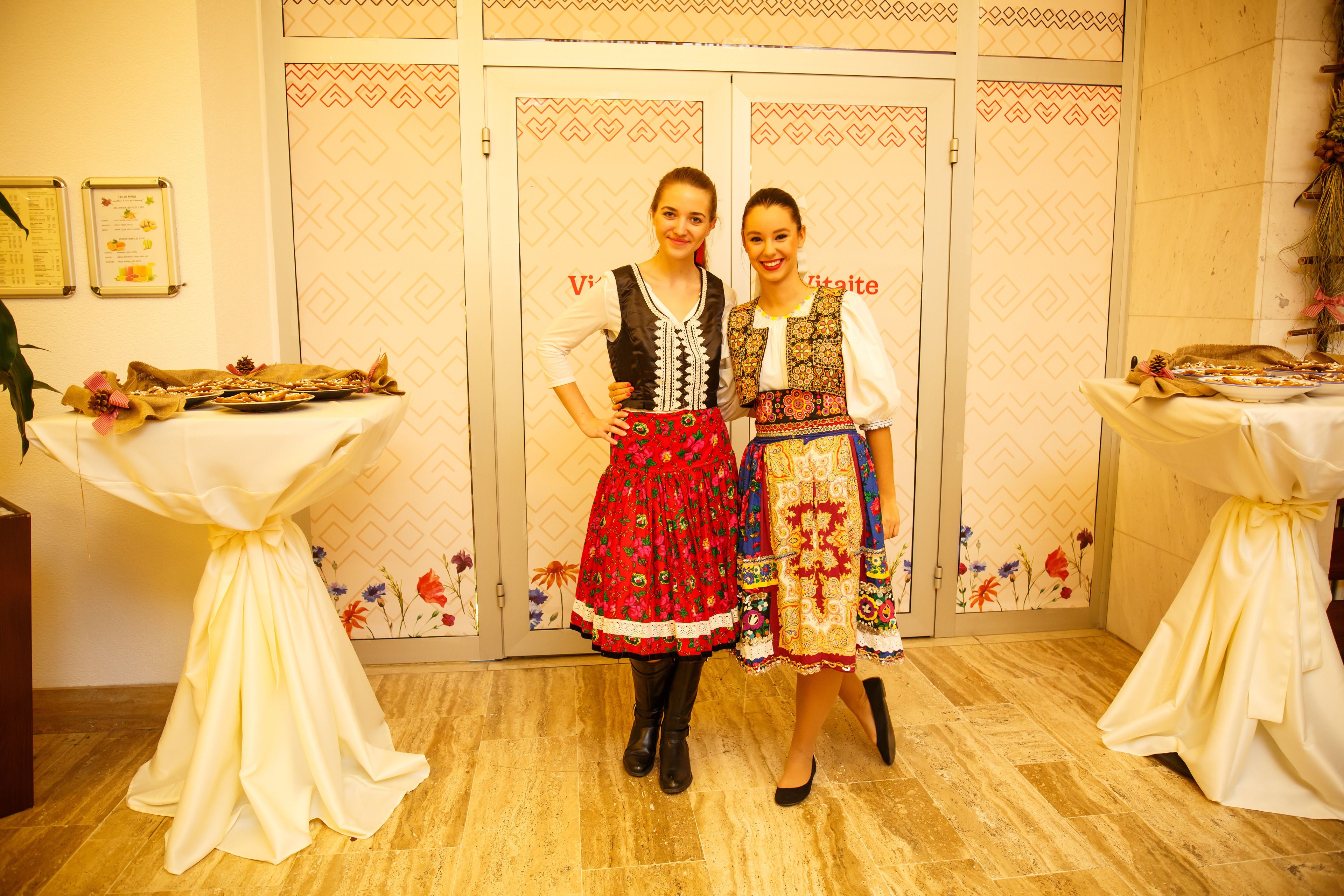 Two women in traditional attire standing in front of a patterned wall with a table displaying food items.
