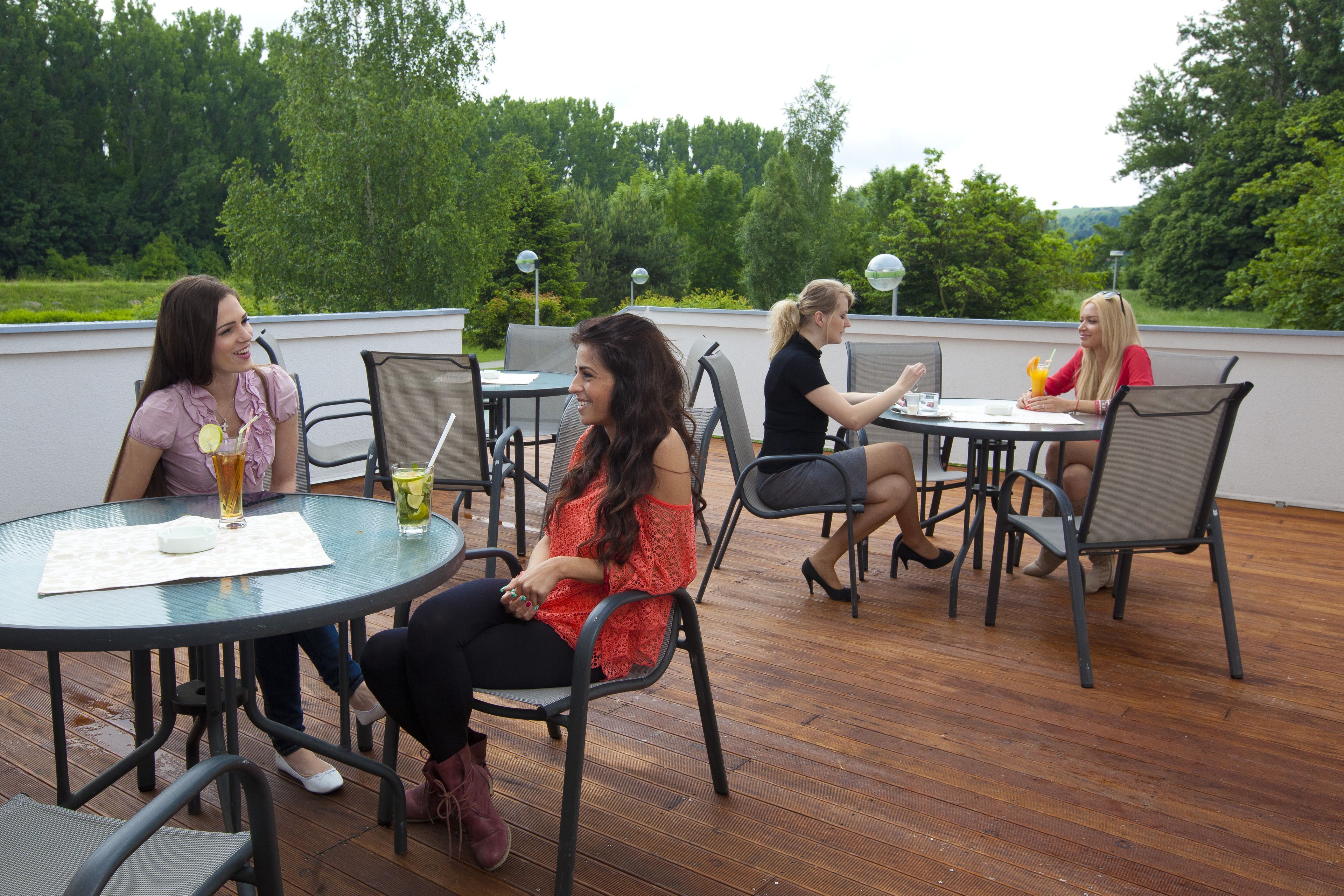 Eine Gruppe von Personen sitzt an runden Tischen auf einer Terrasse mit Holzboden und Metallstühlen.