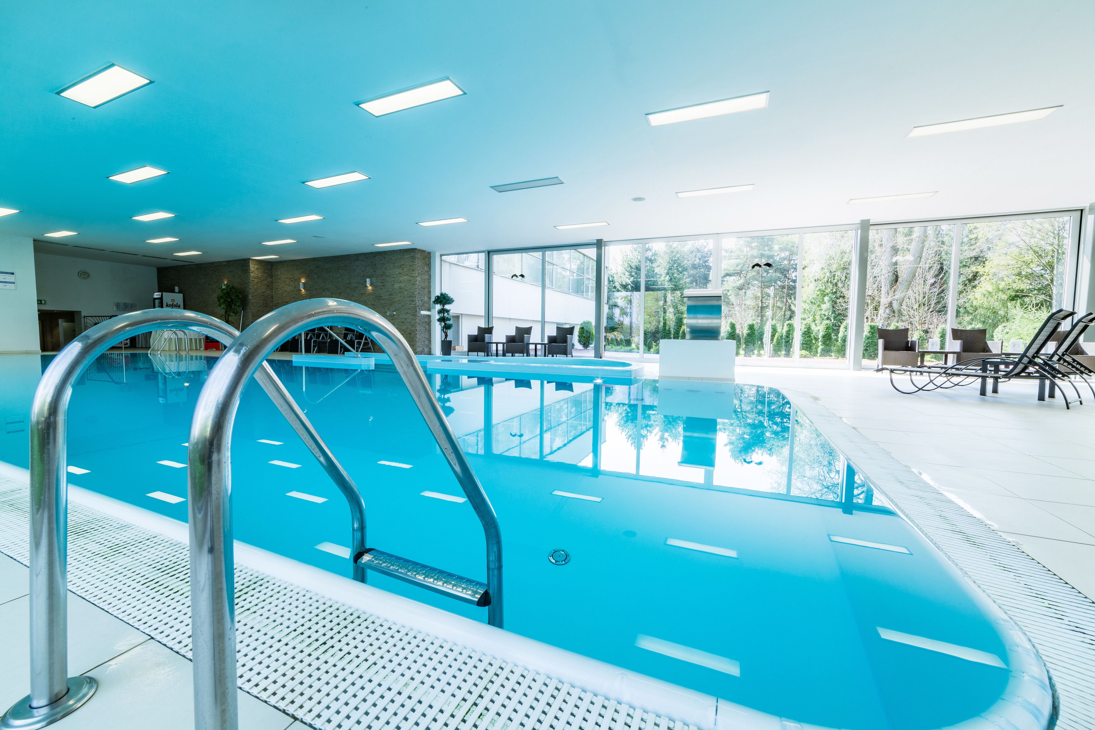 An indoor swimming pool with clear blue water, metal pool ladders, and a seating area with chairs and tables.
