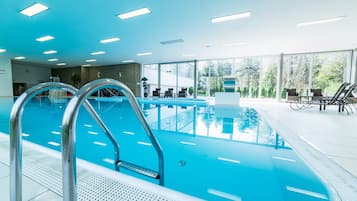 An indoor swimming pool with clear blue water, metal pool ladders, and a seating area with chairs and tables.