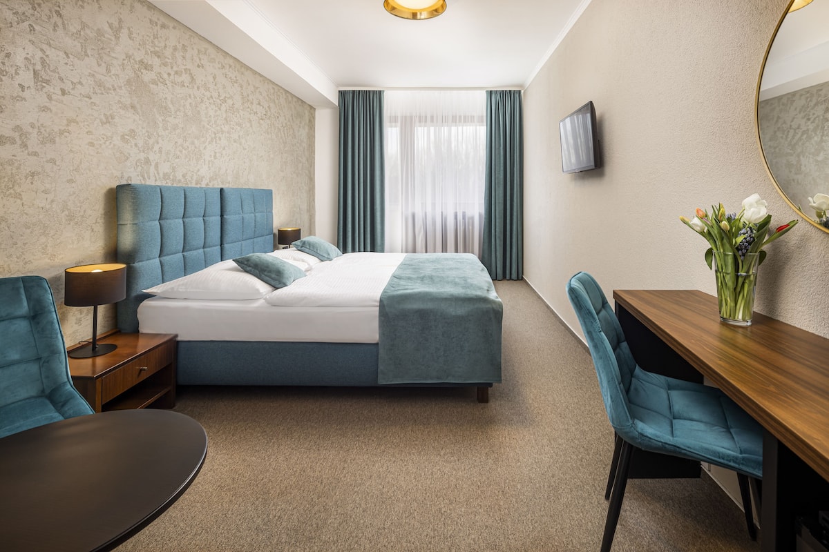 A modern hotel room with a large bed, a desk, and a chair. There is a TV on the wall and a window with curtains.