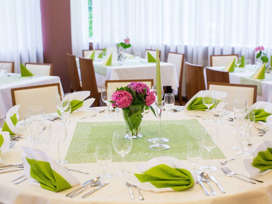 A dining area with round tables set for a formal meal, each with a green table runner, white napkins, and a vase of pink flowers.