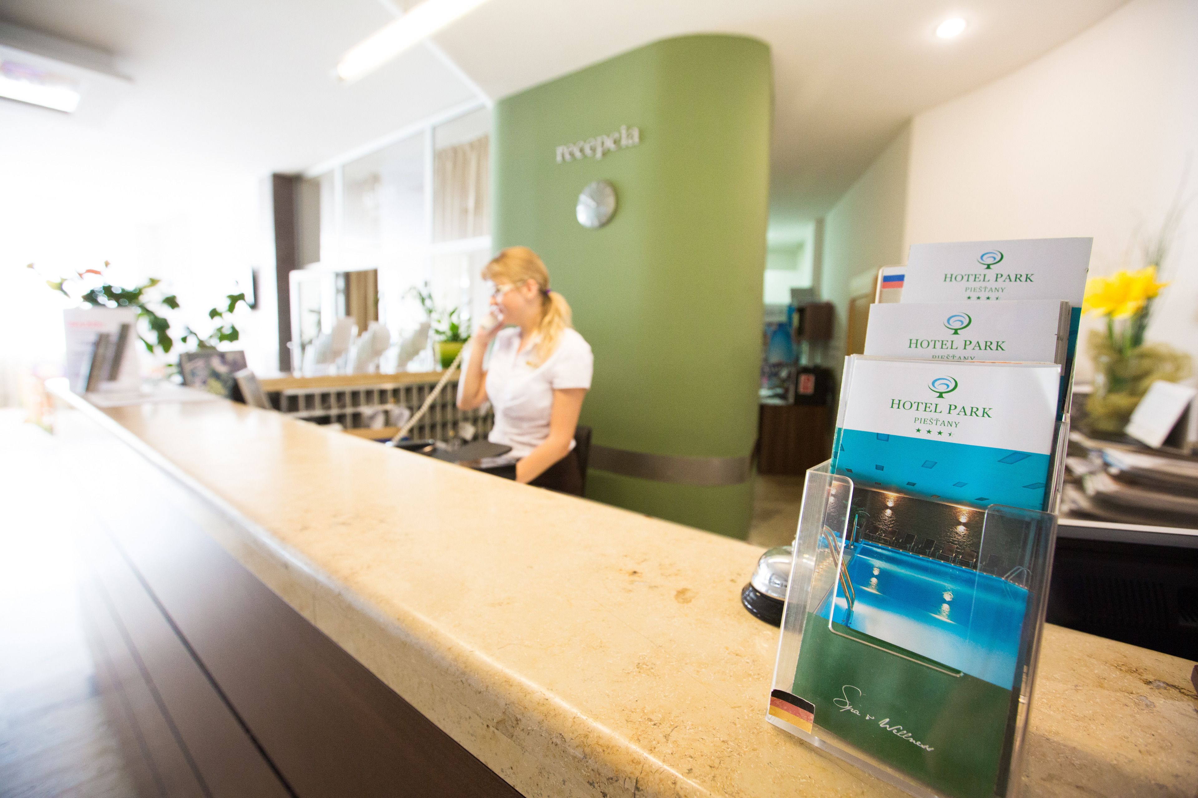 Hotellin vastaanottotiski, jonka takana on vihreä seinä ja seinäkello. Tiskillä on esitteitä Hotel Park Piestany -hotellista.