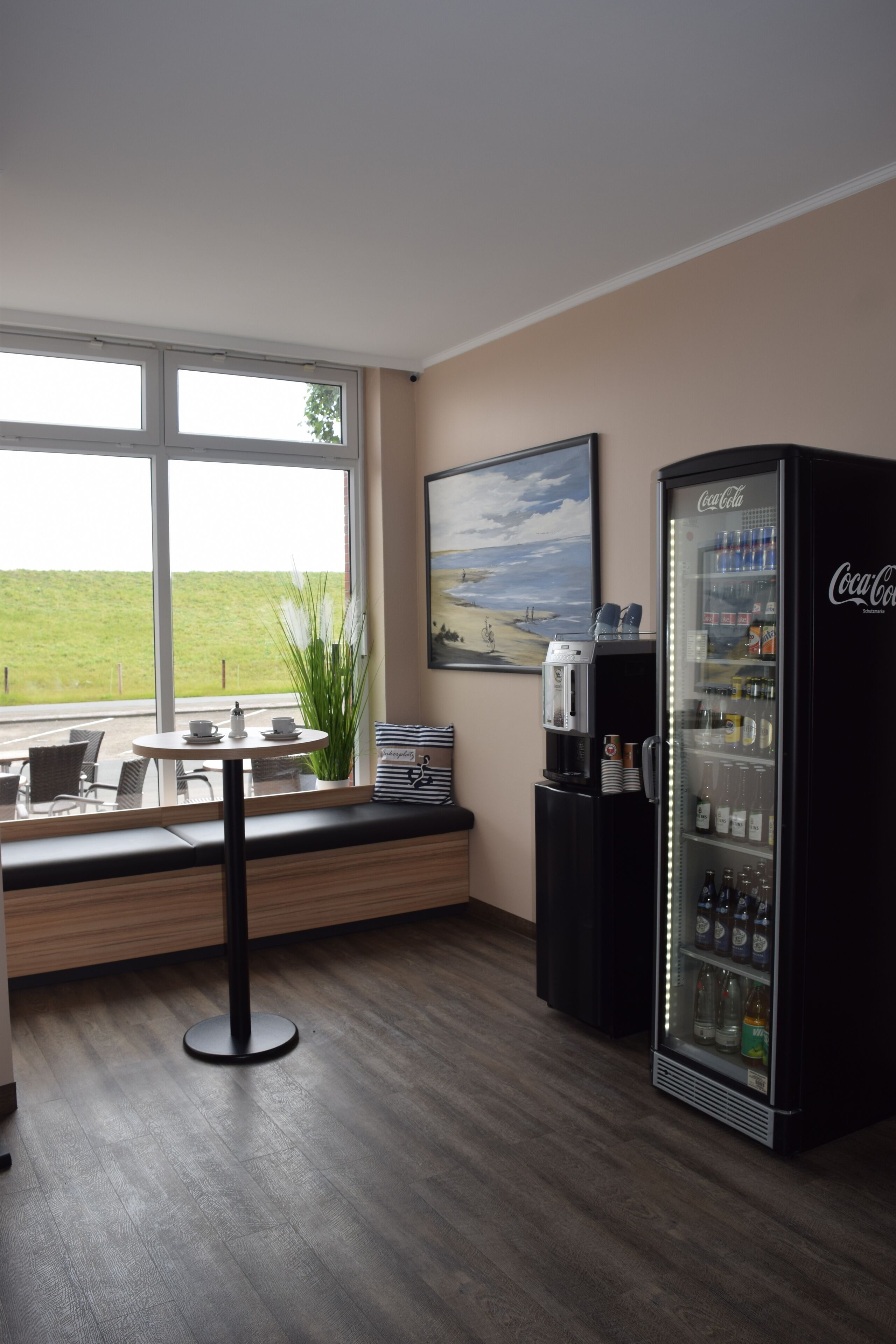 A room with a large window, a seating area with a table and chairs, a Coca-Cola refrigerator, and a painting on the wall.