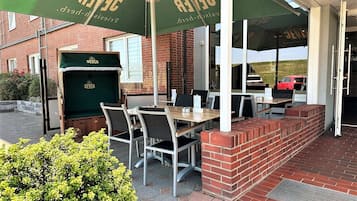 Un espace extérieur aménagé avec des tables et des chaises sous des parasols verts, un mur de briques et une voiture en arrière-plan.