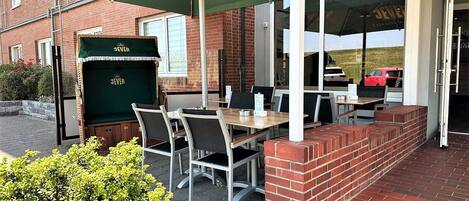 An outdoor seating area with tables and chairs under green umbrellas, a brick wall, and a car in the background.