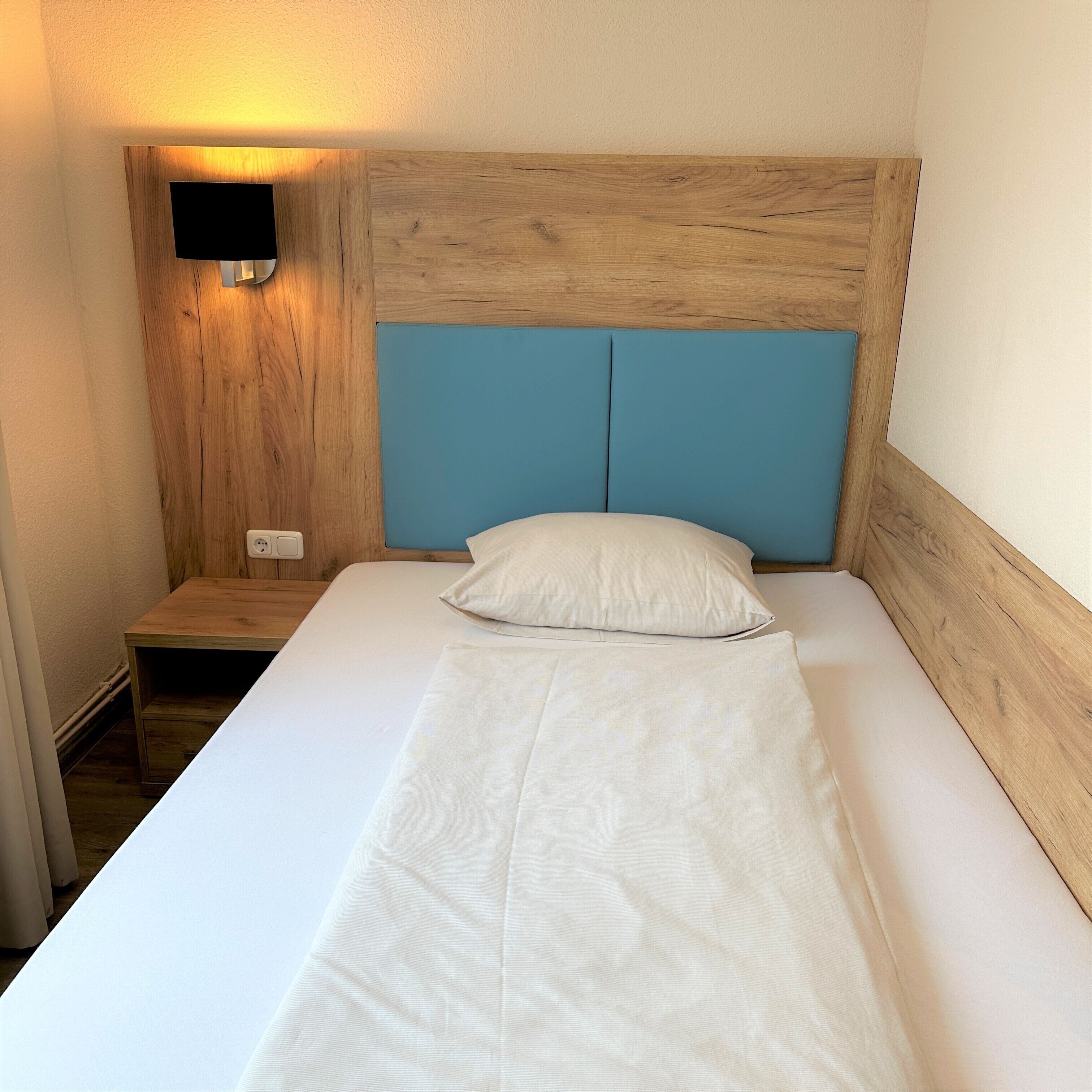 Un lit simple avec une tête de lit en bois et un panneau bleu, une table de chevet et une lampe fixée au mur.