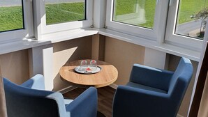 Una habitación de esquina con una mesa redonda de madera y dos sillas azules, ventanas amplias con vistas al mar, y una pequeña mesa con dos vasos y una botella.
