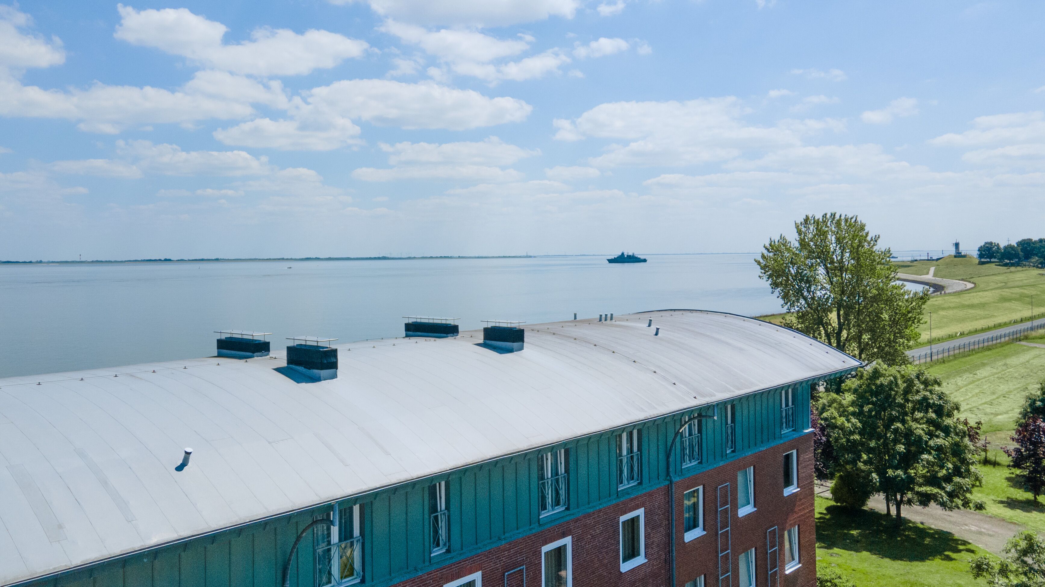 A building with a flat roof overlooking a body of water, a grassy area with a fence, and a distant ship.