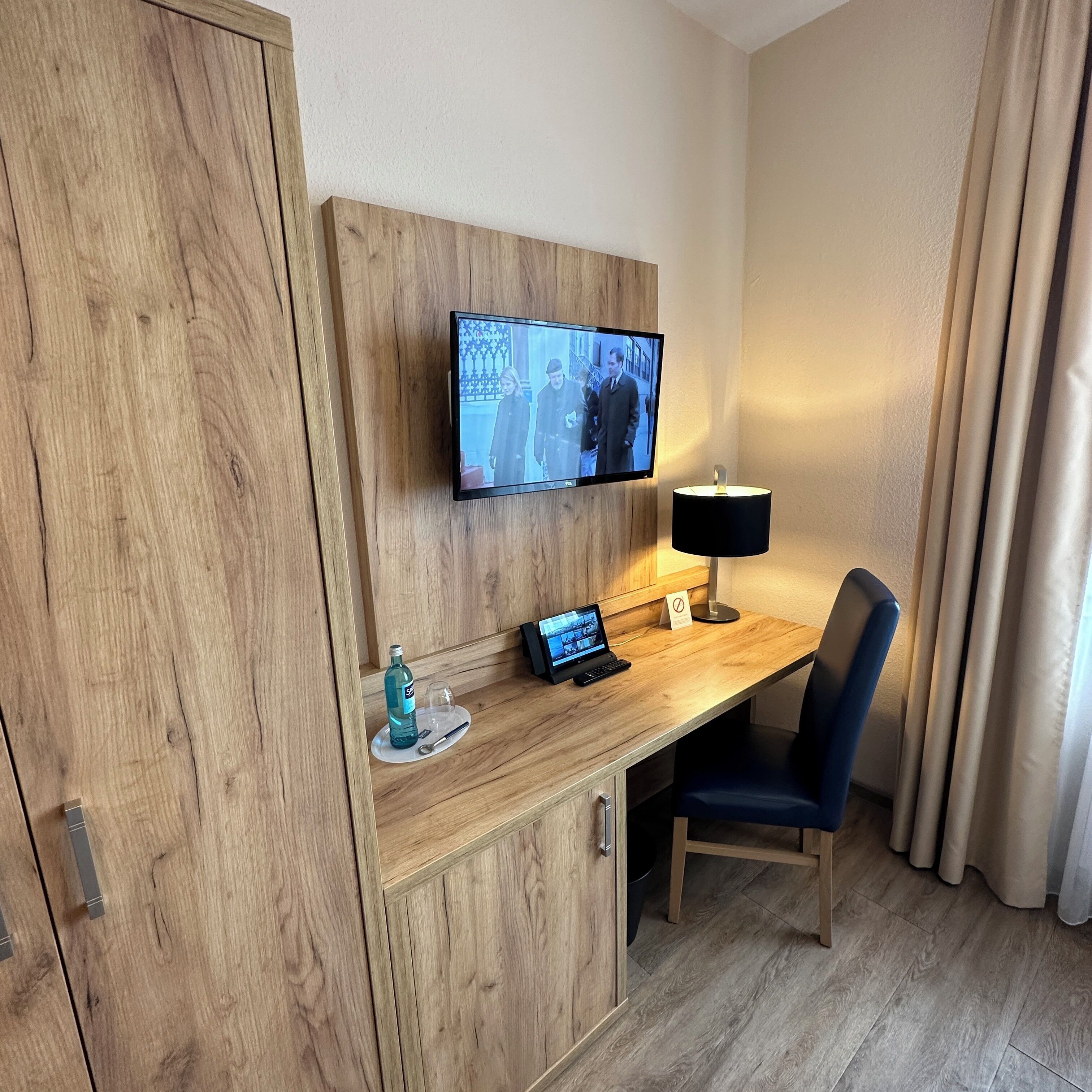A hotel room with a wooden desk, a flat-screen TV mounted on the wall, a desk lamp, a laptop, and a chair.