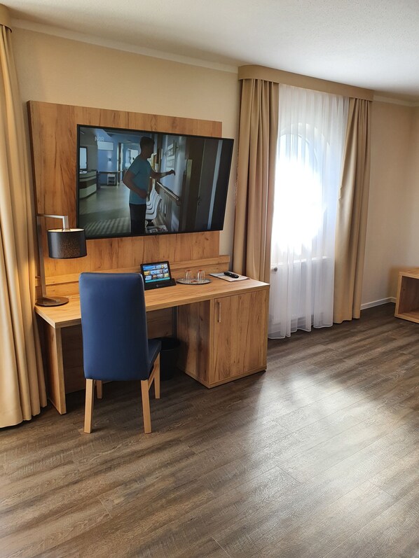 Una habitación de hotel con un escritorio de madera, una silla azul, un televisor montado en la pared y una ventana con cortinas de gasa.