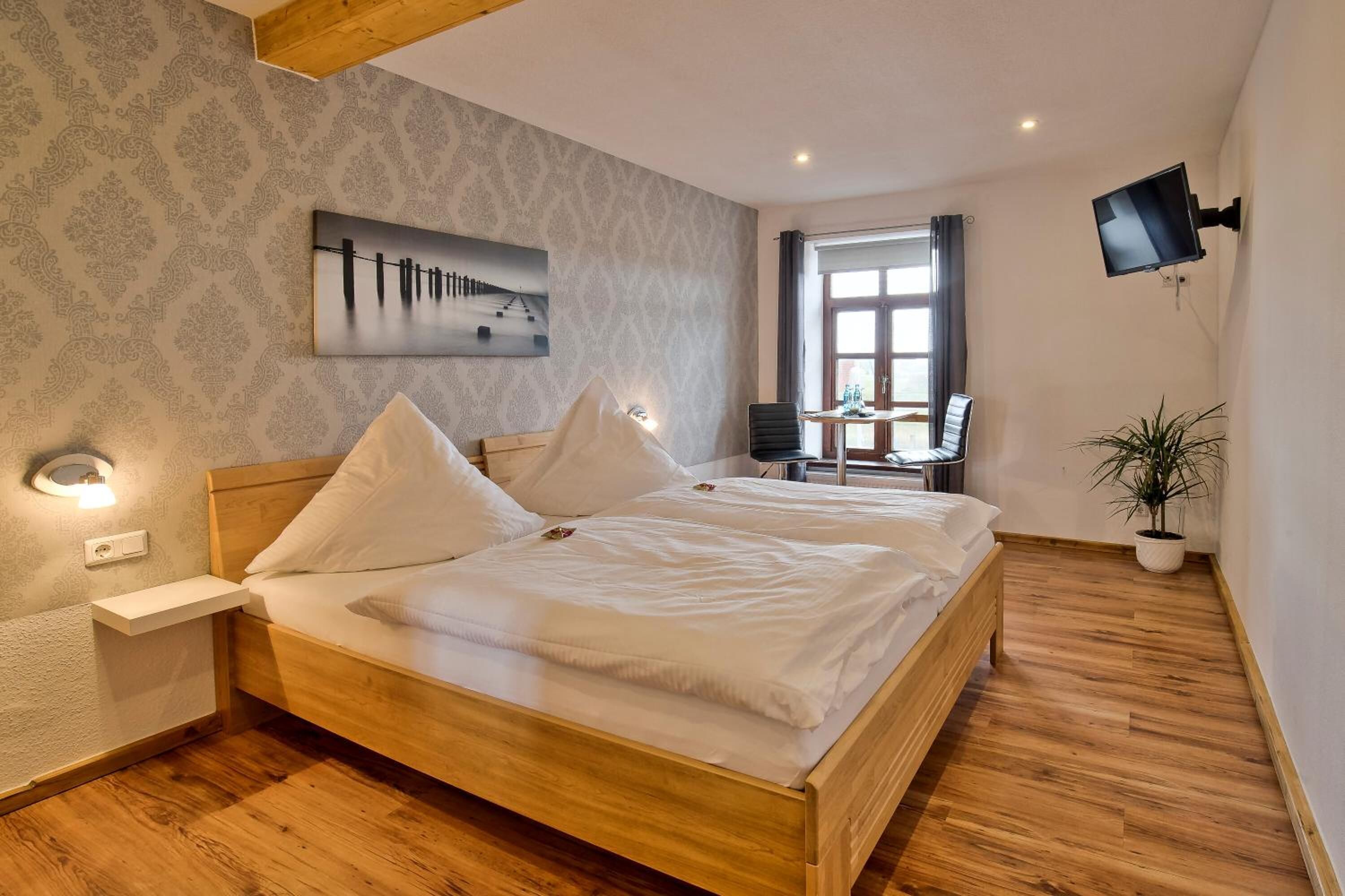 A bedroom with a wooden bed, a nightstand, a desk with a chair, a potted plant, and a painting on the wall.