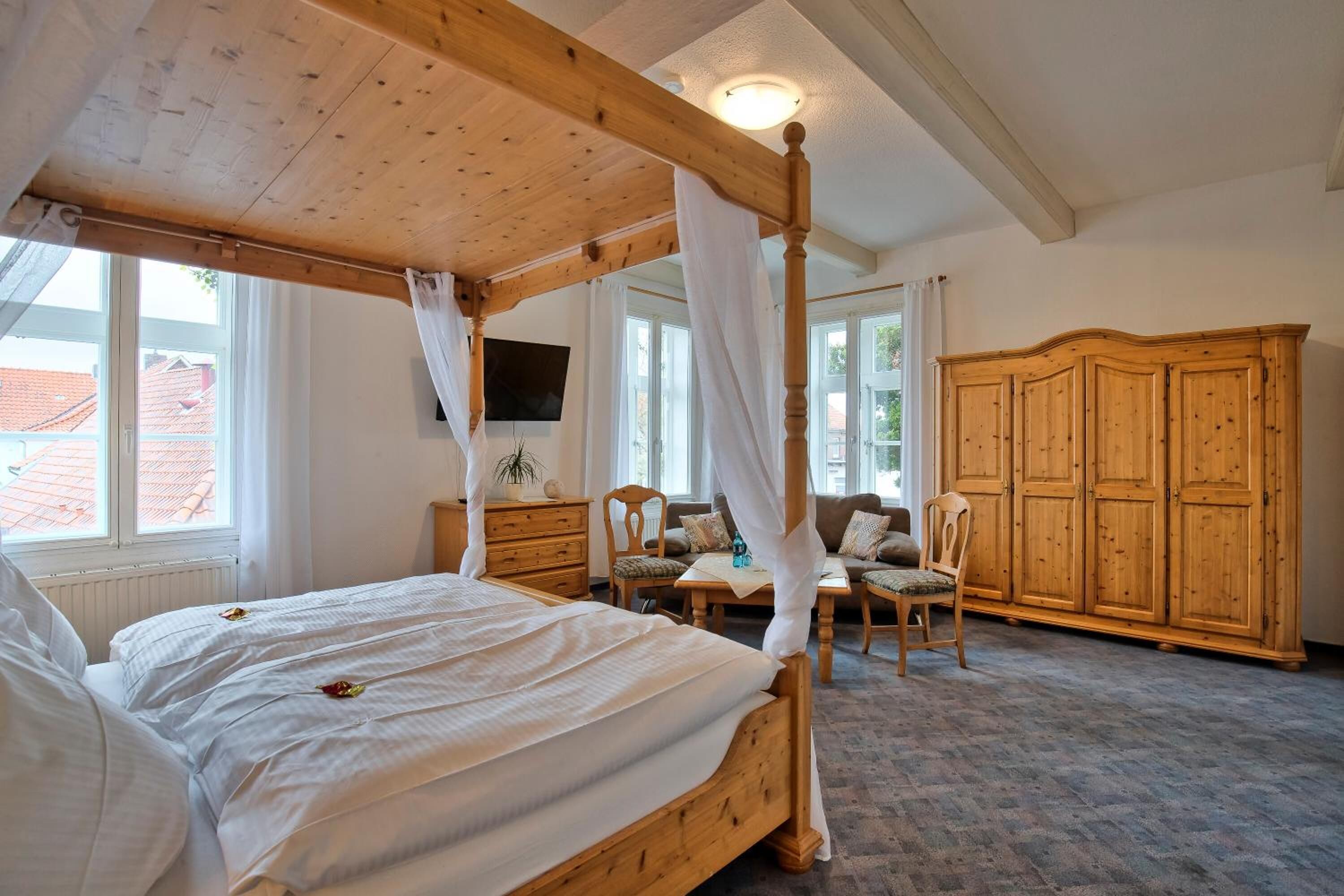A four-poster bed with a canopy, wooden furniture, and a large wardrobe in a room with large windows.