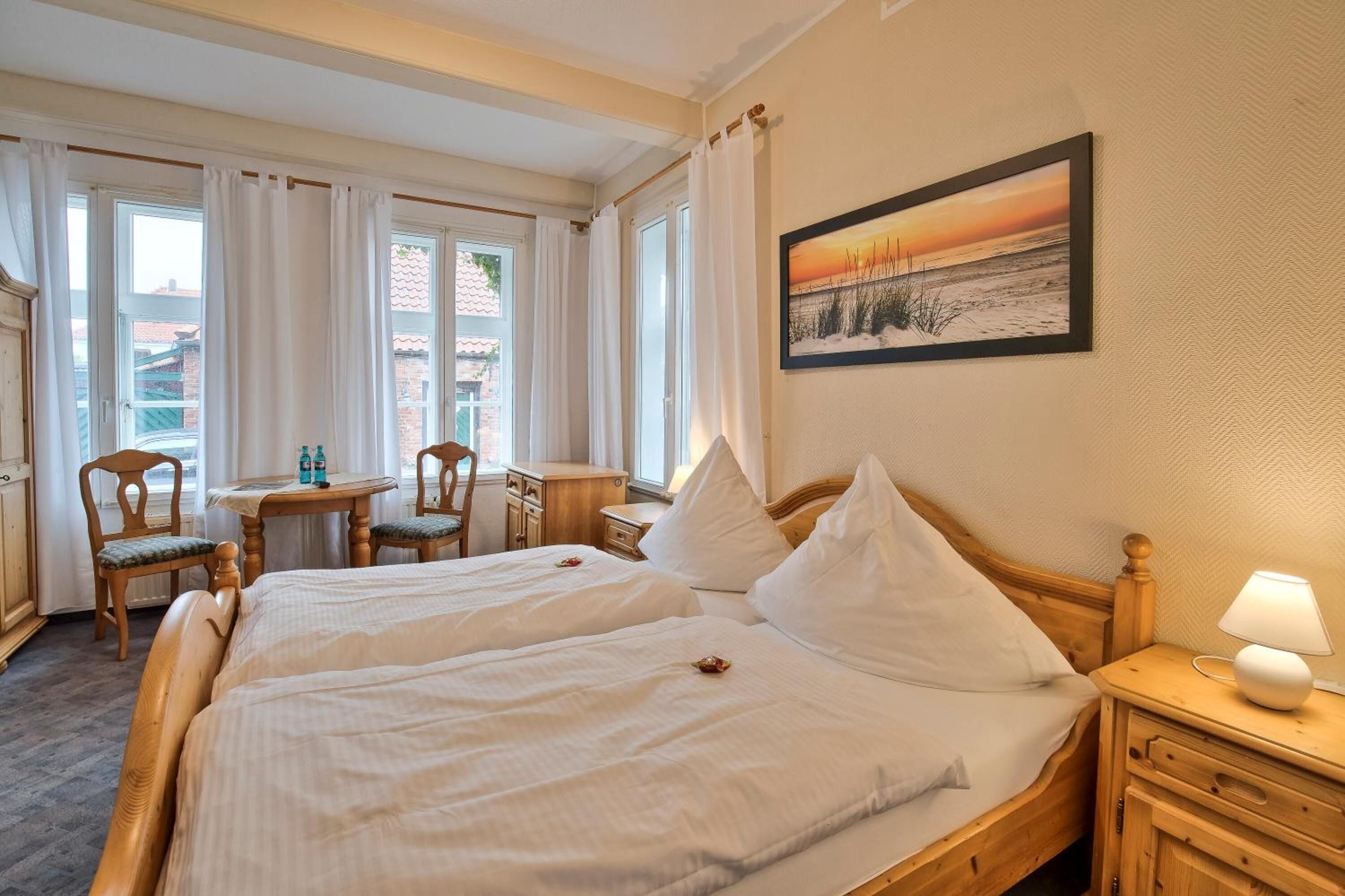 A hotel room with two beds, a small table with chairs, a lamp, and a painting of a sunset.