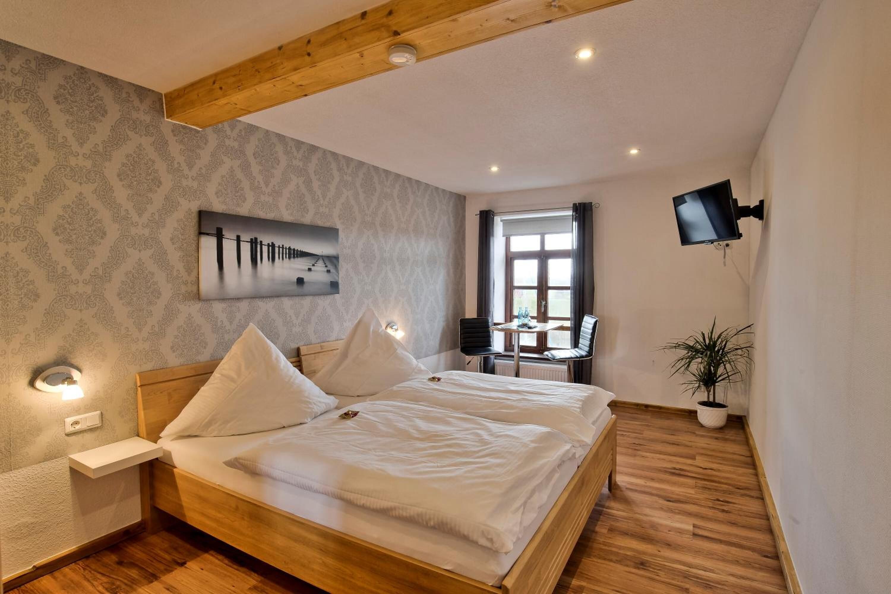 A bedroom with a wooden bed, a nightstand, a wall-mounted TV, a window with curtains, and a potted plant.
