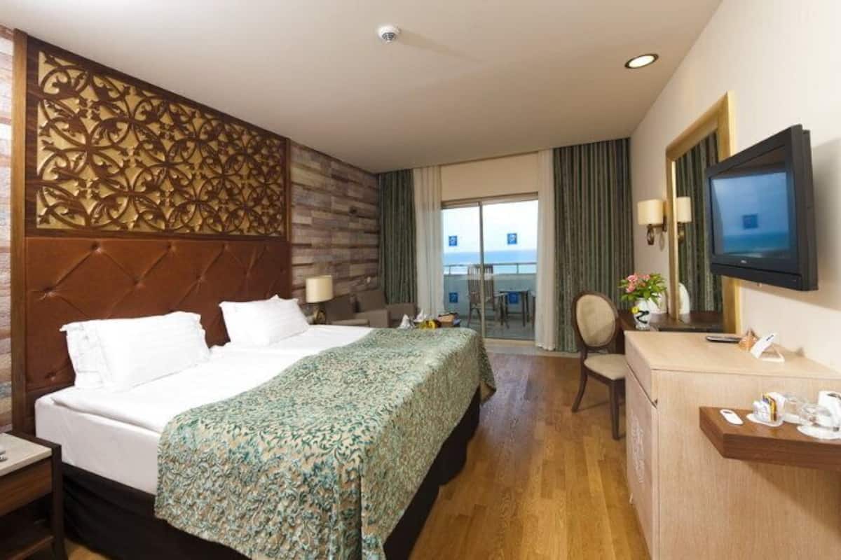 A hotel room with a large bed, a desk with a TV, and a view of the sea through the window.