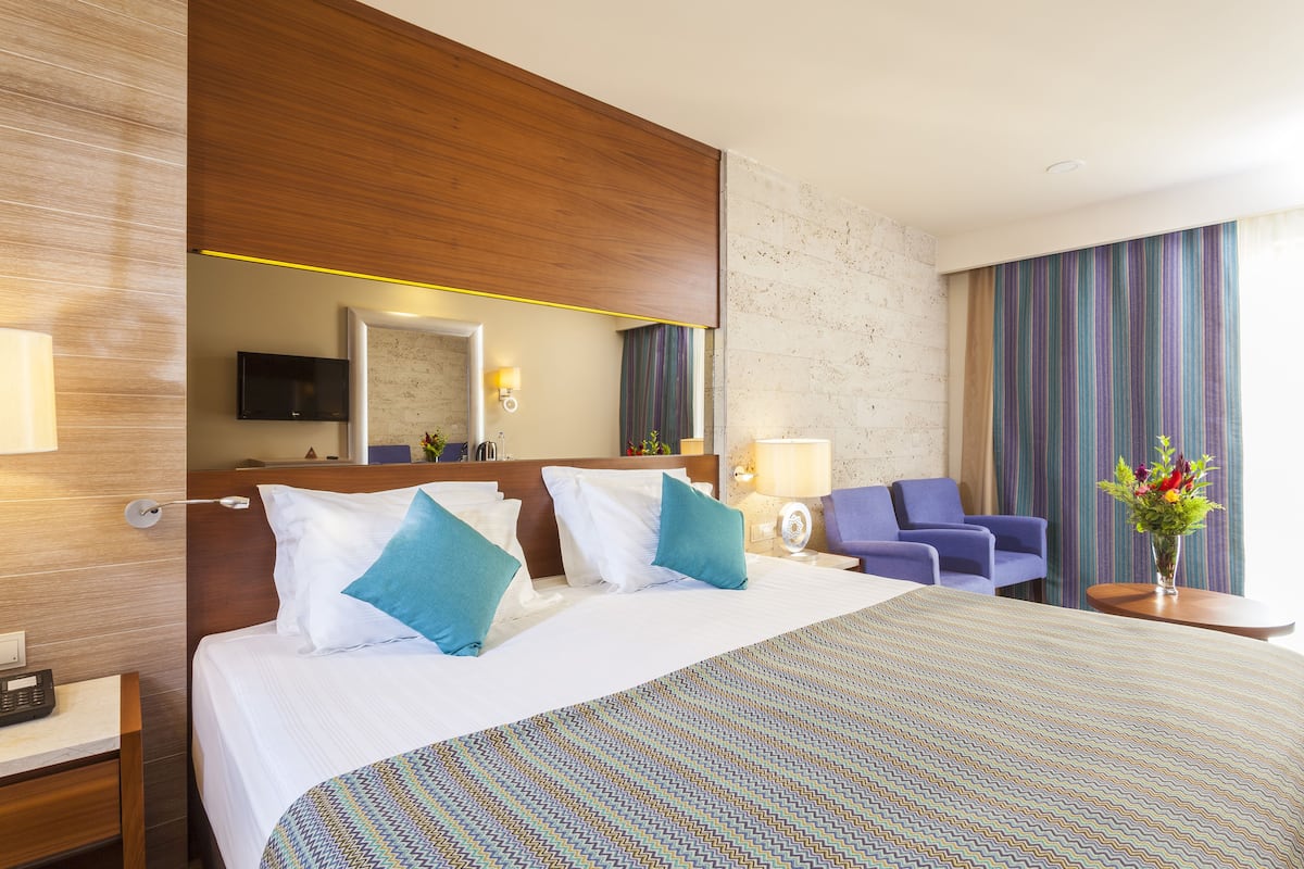A hotel room with a large bed, a wooden headboard, a TV, a desk with a lamp, a chair, and a vase with flowers.