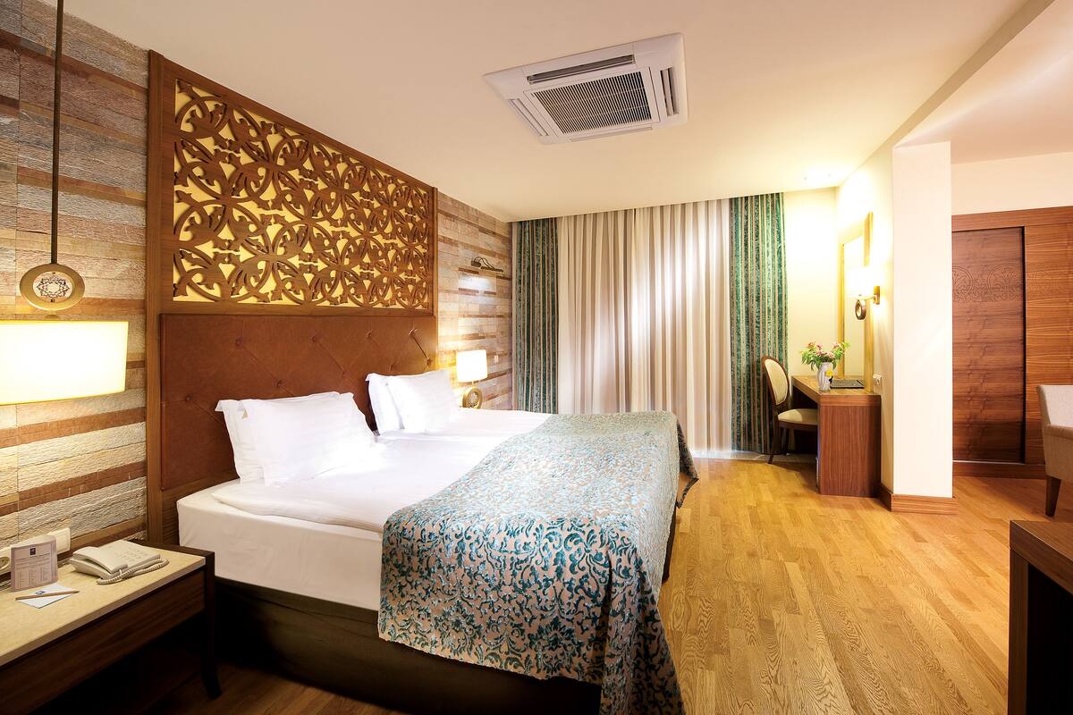 A hotel room with a large bed, a bedside table with a lamp, a desk with a chair, a wooden headboard, and a decorative panel.