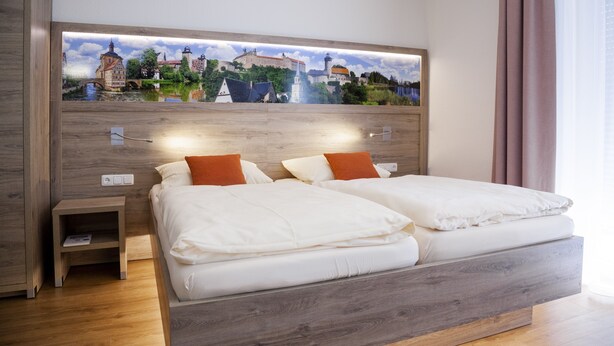 Cama doble con ropa blanca y almohadas naranjas, una mesita de noche con un libro, y un cuadro en la pared que representa un pueblo europeo.