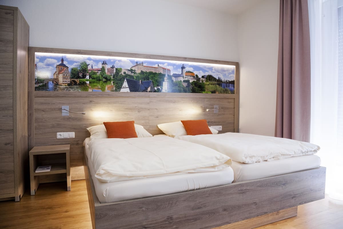 Ein Doppelbett mit weißer Bettwäsche und orangenen Kissen, ein Nachttisch mit einem Buch sowie ein Wandbild einer europäischen Stadt.